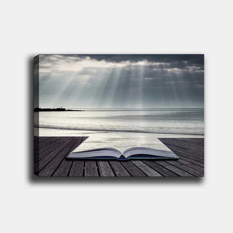 Lerretsbilde 70x100 cm - Åpen bok liggende på en trebrygge ved sjøen, omgitt av myke lysstråler fra en overskyet himmel, Grå / Blå / Hvit