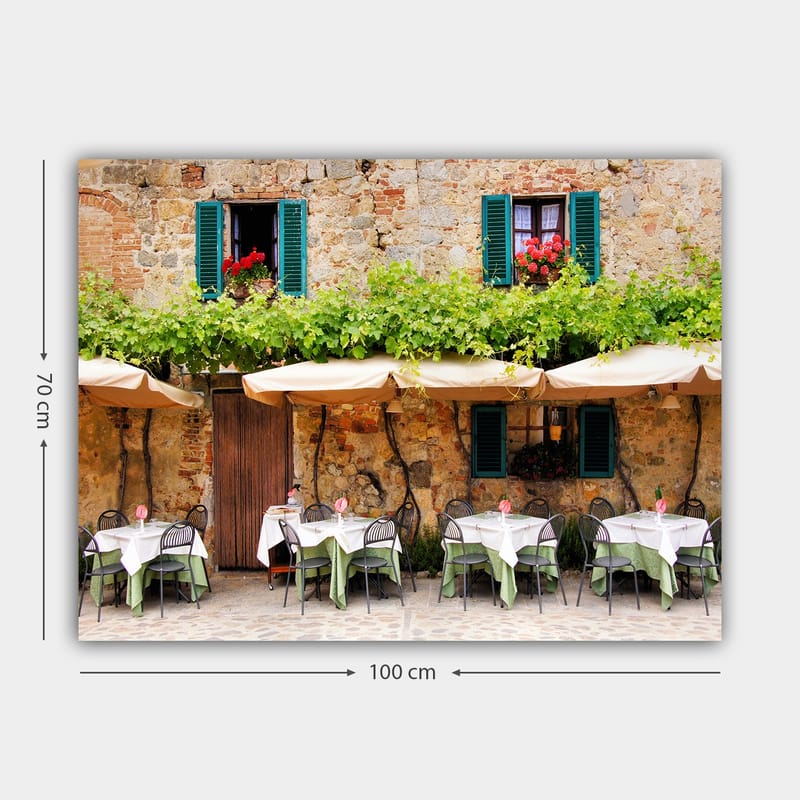 Lerretsbilde 70x100 cm - Sjarmerende utendørs spiseplass med hvite bord og grønne duker foran en gammel steinbygning med mørkegrønne skodder - Mørkegrønn / Hvit / Mursteinsrød - Innredning - Bilder & kunst - Lerretsbilder