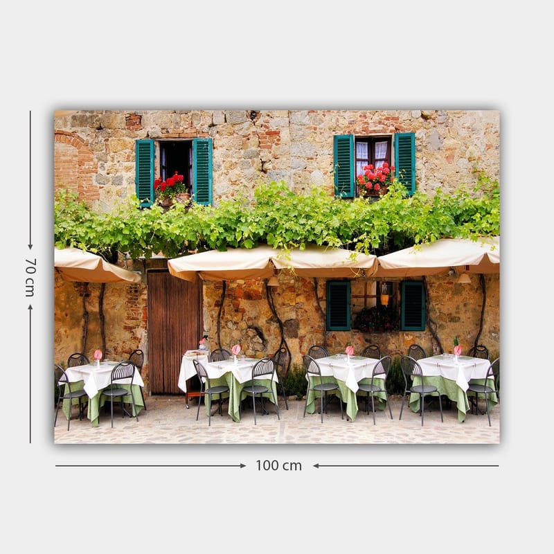 Lerretsbilde 70x100 cm - Sjarmerende utendørs spiseplass med hvite bord og grønne duker foran en gammel steinbygning med mørkegrønne skodder - Mørkegrønn / Hvit / Mursteinsrød - Innredning - Bilder & kunst - Lerretsbilder