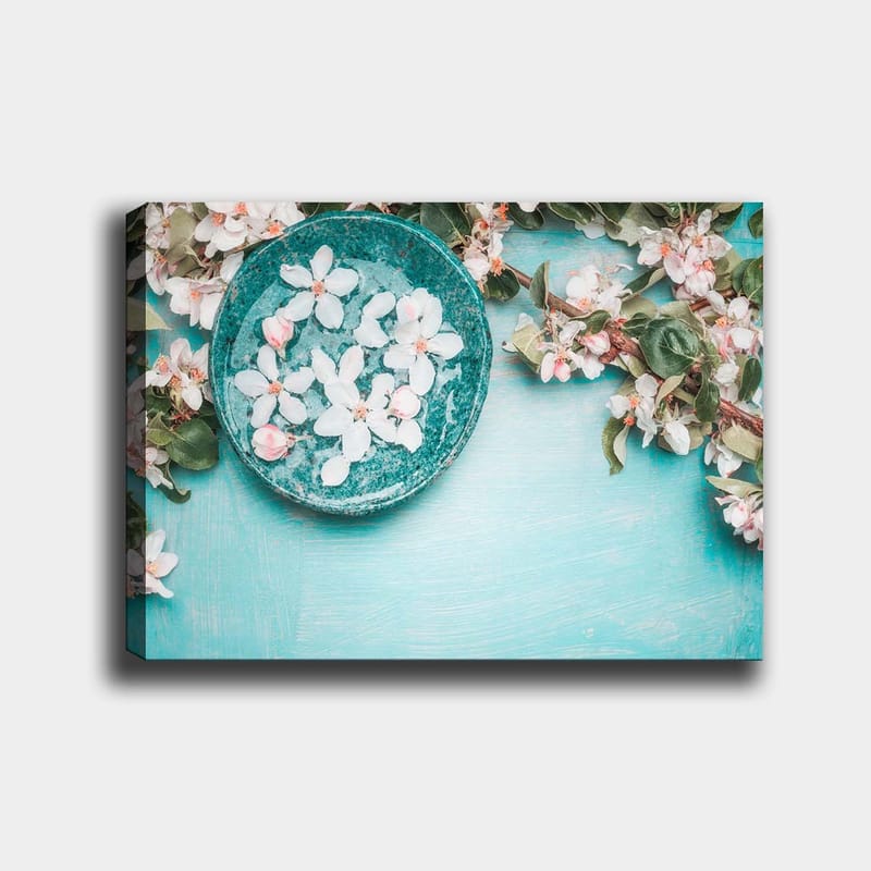 Lerretsbilde 70x100 cm - Skål med blomster omgitt av frodige grønne kvister og blomster på en lyseblå bakgrunn, Turkis / Hvit / Rosa