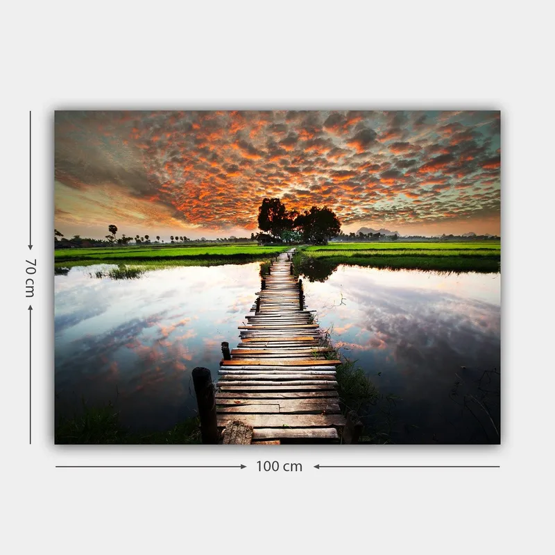 Lerretsbilde 70x100 cm - Trebro som spenner over en speilblank vannoverflate, omgitt av frodig grøntområde og en dramatisk solnedgang - Oransje / Grønn / Blå - Innredning - Bilder & kunst - Lerretsbilder