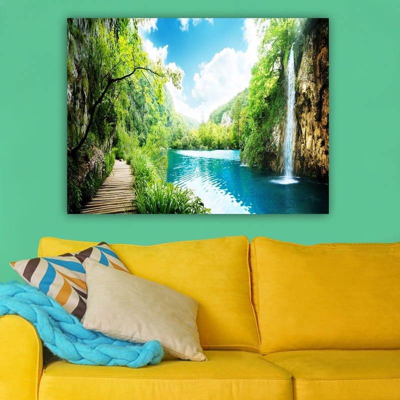 Lerretsbilde 70x100 cm - Vakker naturscene med en foss som renner ut i en klar, blå innsjø omgitt av frodig grøntområde og fjell - Grønn / Blå - Innredning - Bilder & kunst - Lerretsbilder