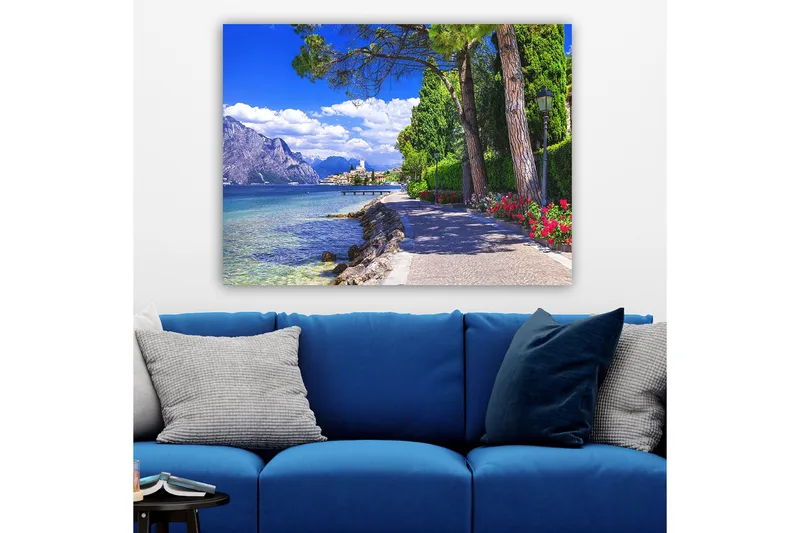 Lerretsbilde 70x100 cm - Vakker promenade med trær og blomster ved en innsjø, omgitt av fjell og en pittoresk by i bakgrunnen - Blå / Grønn / Rød - Innredning - Bilder & kunst - Lerretsbilder