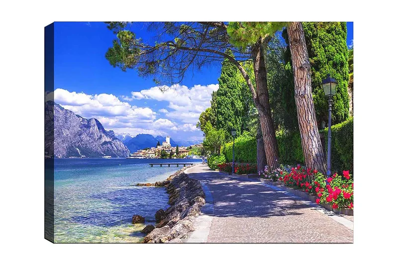 Lerretsbilde 70x100 cm - Vakker promenade med trær og blomster ved en innsjø, omgitt av fjell og en pittoresk by i bakgrunnen, Blå / Grønn / Rød