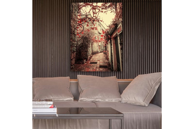 Lerretsbilde 70x100 cm - Pittoresk smug omkranset av gamle bygninger og blomster i røde fargetoner, Rød / Beige