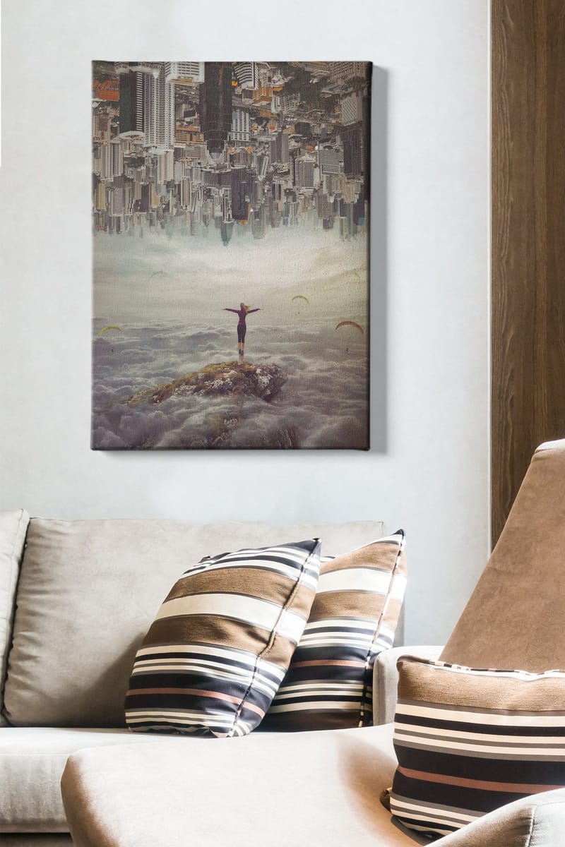 Lerretsbilde 70x100 cm - Person som står med utstrakte armer på en klippe over skyene, med en by i bakgrunnen som er opp ned - Grå / Beige / Mørk blå - Innredning - Bilder & kunst - Lerretsbilder